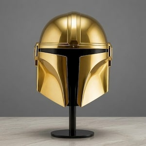 May include: A polished gold-colored helmet featuring a smooth, reflective surface and a distinct T-shaped black visor opening. The helmet is mounted on a simple black cylindrical stand, showcasing a sleek, symmetrical design with curved panels.