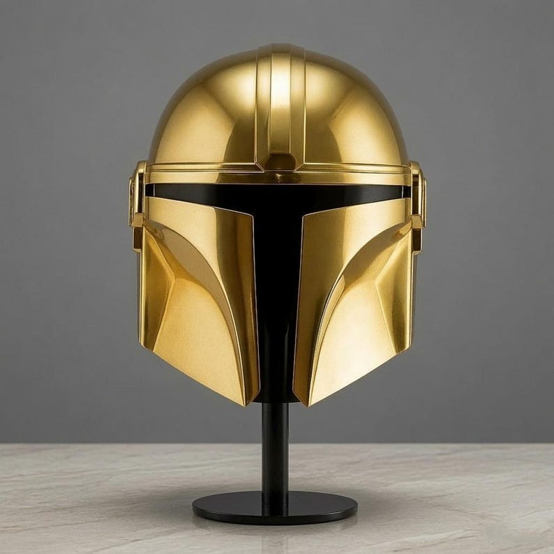 May include: A polished gold-colored helmet featuring a smooth, reflective surface and a distinct T-shaped black visor opening. The helmet is mounted on a simple black cylindrical stand, showcasing a sleek, symmetrical design with curved panels.