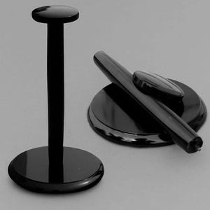 May include: A black display stand with a circular base and a vertical post topped with a small disc, alongside a separate black circular base featuring a slanted cylindrical holder and a small oval-shaped component resting on its surface.