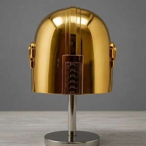May include: A polished gold-colored helmet with a smooth, rounded dome shape and a vertical rectangular panel featuring horizontal ridges on the front. The helmet is mounted on a silver-colored cylindrical stand with a circular base.
