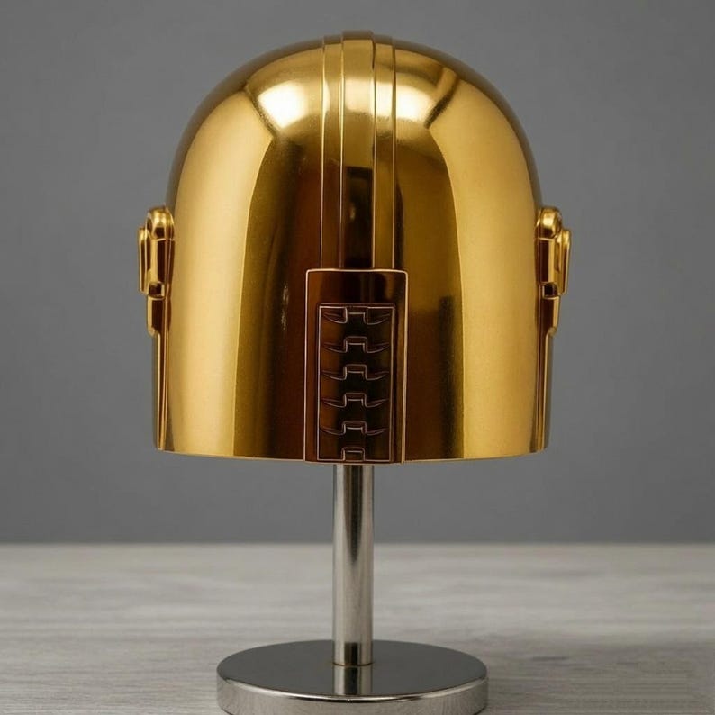 May include: A polished gold-colored helmet with a smooth, rounded dome shape and a vertical rectangular panel featuring horizontal ridges on the front. The helmet is mounted on a silver-colored cylindrical stand with a circular base.