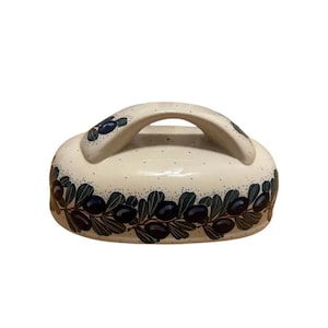 May include: Cream-colored ceramic serving dish with a handle. The dish is decorated with a dark blue olive branch design around the base and on the handle. Small black dots are scattered across the surface.