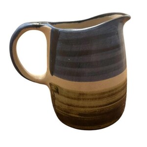 May include: A ceramic pitcher featuring a rounded body and a curved handle. The upper portion is glazed in a dark blue-grey tone, while the lower section displays a natural tan base with horizontal brushstrokes in a muted olive green hue.
