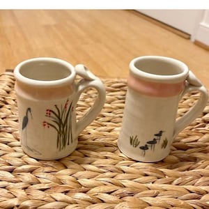 May include: Two ceramic mugs with a cream base and pink rim. One mug features a heron and plant design, the other has a bird and plant design. Both mugs have a curved handle and are set on a woven surface.