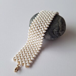 Op de afbeelding: Een witte kralenarmband met goudkleurige accenten en een goudkleurige sluiting. De armband is gedrapeerd over een grijze steen en toont het ingewikkelde patroon van de kralen. De kralen zijn in een geweven ontwerp gerangschikt.