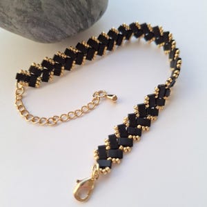 Peut inclure: Un bracelet de perles noir et or avec un motif en zigzag. Le bracelet présente de petites perles noires rectangulaires et des accents dorés. Il possède une chaîne et un fermoir dorés.