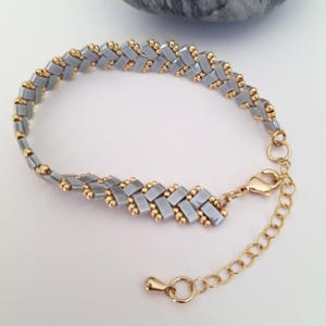 Op de afbeelding: Handgemaakte armband met een chevronpatroon. De armband is gemaakt van kleine, rechthoekige, lichtgrijze kralen en goudkleurige accenten. Het heeft een goudkleurige sluiting en een verstelbare ketting.