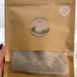 Puede incluir: Una bolsa de papel marrón con una ventana transparente, que contiene té de baño botánico. La bolsa tiene una etiqueta redonda con el texto "Murmure de la nature" y "Whisper of Nature". La bolsa está sellada y tiene una pegatina "Made in Canada".