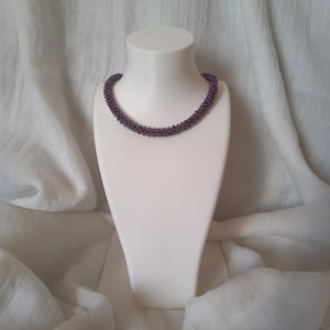 Può includere: Una collana di perline viola scuro con un design tubolare intrecciato. La collana è composta da numerose piccole perline uniformi strettamente collegate tra loro per formare un filo continuo e arrotondato che poggia uniformemente.