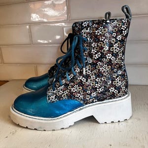 May include: A lace-up ankle boot featuring a metallic blue toe and heel counter, contrasted with a black upper panel covered in a dense white, yellow, and blue floral pattern. The boot is finished with a thick, white, lugged rubber sole.