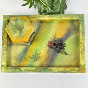 May include: A rectangular resin tray with a matching hexagonal box. The tray features a cannabis leaf and a skull with a rose design. The tray and box have a green, yellow, and gray color scheme.