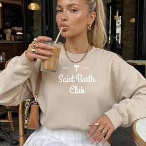 Puede incluir: Sudadera beige con el texto blanco "Saint Barth Club" y un gr&aacute;fico de palmera. Se combina con una falda plisada blanca. La imagen tambi&eacute;n muestra joyas doradas y una bebida en un vaso.