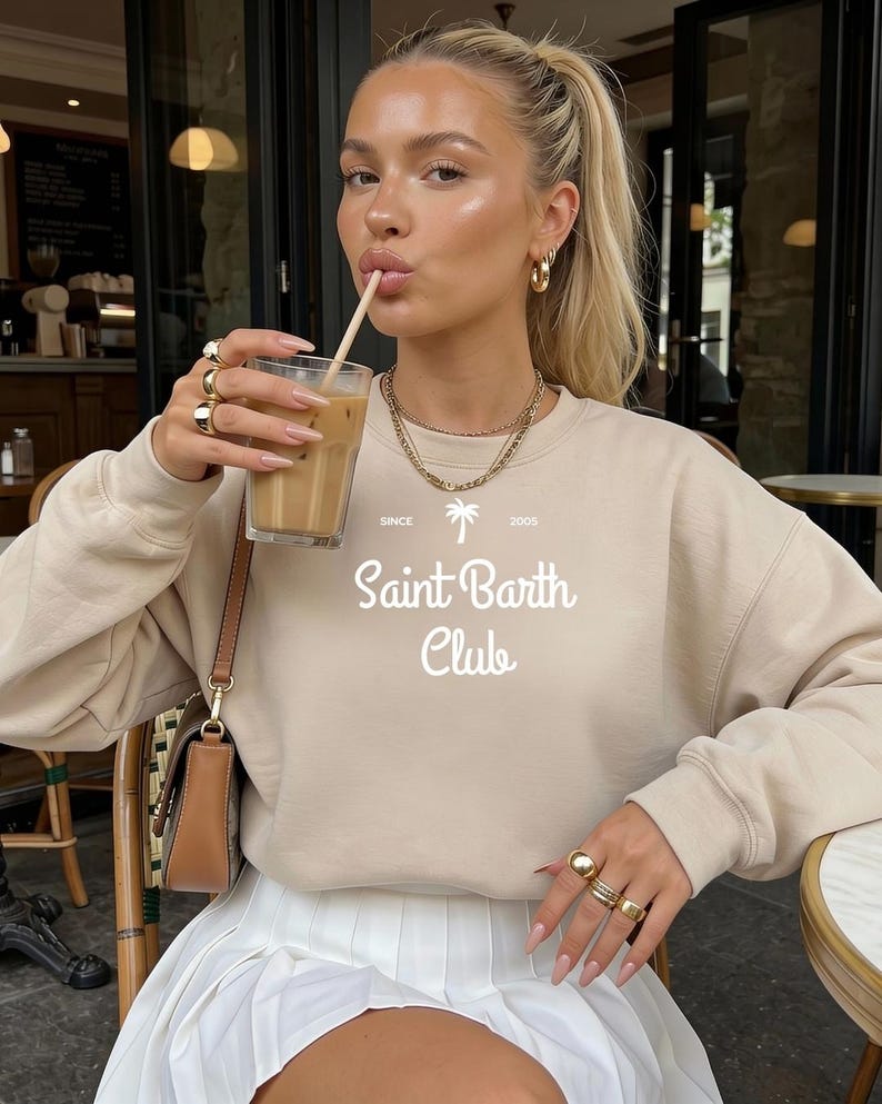 Puede incluir: Sudadera beige con el texto blanco "Saint Barth Club" y un gr&aacute;fico de palmera. Se combina con una falda plisada blanca. La imagen tambi&eacute;n muestra joyas doradas y una bebida en un vaso.