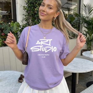 以下が含まれることがあります： 白文字で「ART STUDIO」と「CREATIVE PEOPLE CLUB」がプリントされた、薄紫色のTシャツ。白いプリーツスカートと合わせています。小さな茶色のハンドバッグとサングラスも写っています。