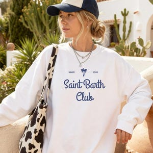 Puede incluir: Sudadera blanca con el texto "Saint Barth Club" en azul marino, junto con un gr&aacute;fico de palmera. El conjunto incluye una gorra de b&eacute;isbol azul marino y crema y un bolso tote con estampado de vaca en blanco y negro.