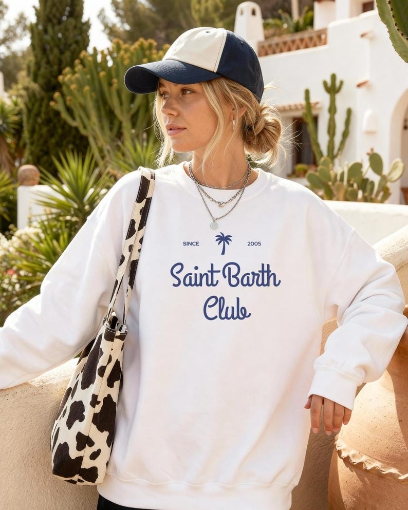 Puede incluir: Sudadera blanca con el texto "Saint Barth Club" en azul marino, junto con un gr&aacute;fico de palmera. El conjunto incluye una gorra de b&eacute;isbol azul marino y crema y un bolso tote con estampado de vaca en blanco y negro.