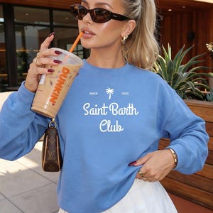 Puede incluir: Una sudadera azul claro con el texto blanco "Saint Barth Club" y un dise&ntilde;o de palmera. La persona lleva gafas de sol y sostiene un caf&eacute; helado Dunkin'. Se ven anillos y una pulsera dorados.