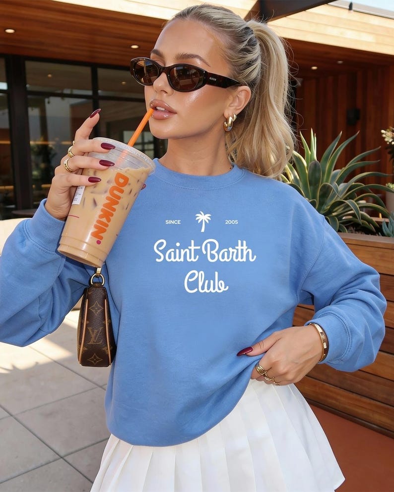 Puede incluir: Una sudadera azul claro con el texto blanco "Saint Barth Club" y un dise&ntilde;o de palmera. La persona lleva gafas de sol y sostiene un caf&eacute; helado Dunkin'. Se ven anillos y una pulsera dorados.