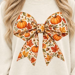 Könnte beinhalten: Ein cremefarbenes Sweatshirt mit Rundhalsausschnitt und einer großen, mittigen Grafik in Form einer Schleife. Die Schleife ist mit einem herbstlichen Muster aus orangefarbenen Kürbissen, braunen Eicheln, roten und orangefarbenen Ahornblättern und kleinen weißen Blüten bedruckt.