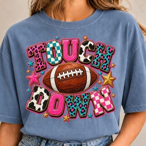 Könnte beinhalten: Ein blaues T-Shirt mit kurzen Ärmeln und einem Grafikdruck mit dem Text TOUCH DOWN. Die Buchstaben sind mit verschiedenen Mustern gefüllt, darunter rosa Leopardenmuster, blau-weiße Karos, Kuhflecken und ein blaues Leopardenmuster.