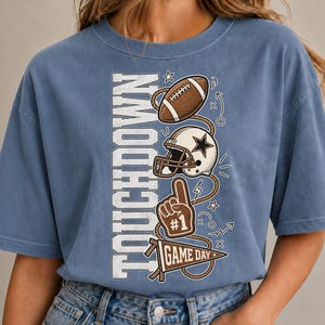 Könnte beinhalten: Ein blaues T-Shirt mit kurzen Ärmeln und einem vertikalen Aufdruck des Wortes TOUCHDOWN in weißen Buchstaben. Das Design zeigt einen braunen Football, einen weißen Helm mit Stern, einen braunen Schaumstofffinger und einen GAME DAY Wimpel.