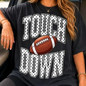 Könnte beinhalten: Ein schwarzes Kurzarm-T-Shirt mit einem großen Grafikdruck, der den Text TOUCH DOWN in weißen Blockbuchstaben mit schwarzen Punkten zeigt. Zwischen den beiden Wörtern befindet sich ein brauner Football mit weißen Schnürsenkeln.
