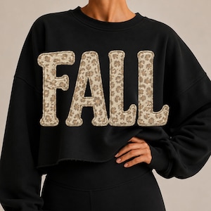 Könnte beinhalten: Ein schwarzes, langärmeliges, bauchfreies Sweatshirt mit dem Wort FALL in großen Blockbuchstaben auf der Brust. Die Buchstaben haben ein beiges und braunes Leopardenmuster mit einer sichtbaren Nahtkante um jeden einzelnen Buchstaben.