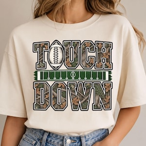 Könnte beinhalten: Ein cremefarbenes T-Shirt mit einem Aufdruck des Textes TOUCH DOWN. Die Buchstaben sind mit einem Tarnmuster aus Baumrinde und Blättern gefüllt. Ein Football ersetzt den Buchstaben O und eine grüne Footballfeld-Grafik liegt zwischen den Wörtern.