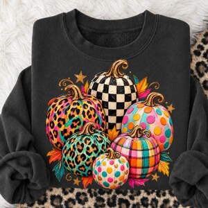 Könnte beinhalten: Ein schwarzes Sweatshirt mit Rundhalsausschnitt und einem Grafikdruck von fünf Kürbissen. Die Kürbisse zeigen verschiedene Muster, darunter schwarz-weiße Karos, buntes Leopardenmuster, leuchtende Punkte und ein mehrfarbiges Karomuster mit goldenen Stielen.
