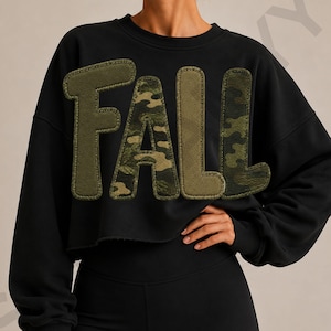 May include: A black long-sleeve cropped sweatshirt featuring the word FALL in large, textured appliqué letters. The letters alternate between a solid olive green fabric and a green, brown, and black camouflage pattern with stitched edges.