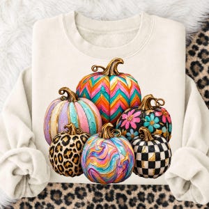 Könnte beinhalten: Ein cremefarbenes Sweatshirt mit Rundhalsausschnitt und einem Grafikdruck von sechs Kürbissen. Die Kürbisse zeigen verschiedene Muster, darunter ein buntes Zickzackmuster, Pastellstreifen, Leopardenmuster, Blumenmotive, eine Wirbeloptik und ein Schachbrettmuster.