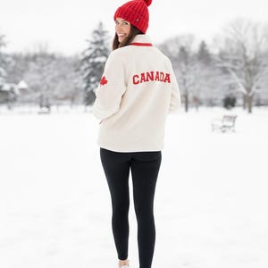 Peut inclure: Une personne porte un bonnet rouge à pompon et une veste polaire crème avec "CANADA" en lettres rouges au dos. La veste a un écusson de feuille d'érable rouge et une bordure rouge. Des leggings noirs et des baskets blanches complètent la tenue.