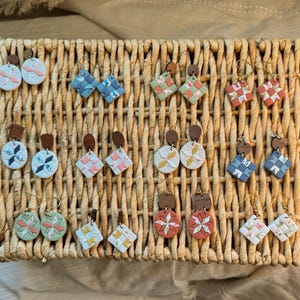 May include: A collection of handmade earrings displayed in a woven basket. The earrings feature various geometric and floral designs in pastel colors, with wooden and gold-tone metal accents. The earrings are a mix of round and square shapes.