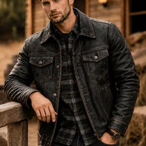 May include: A distressed black leather jacket with a pointed collar, front button closure, and two chest pockets. The jacket is worn over a dark grey and black plaid button-down shirt and dark denim jeans, with a wristwatch visible on the wrist.