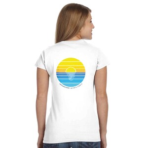 May include: A white short-sleeve t-shirt featuring a circular graphic on the back. The design displays a yellow sun setting over blue ocean waves with horizontal stripes. Below the circle, the text reads PORTHMEOR SUNSET / 50 12 56 N / 5 29 55 W.
