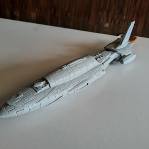 May include: A gray spaceship model with detailed paneling and a fin. The model has a long, streamlined body with a pointed front and a rear fin with orange accents. It is a collectible item.