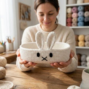 May include: An oval-shaped, off-white crochet basket featuring two upright bunny ears on the rim. The front of the basket displays a simple embroidered face with two black oval eyes and a black stitched cross for a nose and mouth.