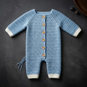 Può includere: Un pagliaccetto per neonati all'uncinetto blu chiaro con maniche lunghe e bottoni in legno sul davanti. I polsini e le caviglie sono rifiniti con il bianco. Il pagliaccetto ha uno scollo tondo e un motivo a punto strutturato.
