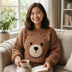 Könnte beinhalten: Ein brauner Häkelpullover mit einem Bärengesicht auf der Vorderseite. Der Bär hat eine beige Schnauze, schwarze Augen und eine schwarze Nase. Der Pullover wird von einer Person getragen, die auf einem Sofa sitzt und eine Tasse Tee und ein Buch hält.
