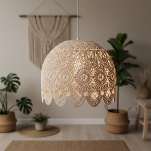 Può includere: Una lampada a sospensione all'uncinetto beige con un bordo smerlato pende dal soffitto. La luce ha una forma rotonda e un motivo a trama aperta e dettagliata. Lo sfondo comprende piante in vasi intrecciati e un arazzo macramè.
