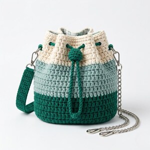 May include: A crochet bucket bag featuring a color-blocked design in cream, light green, and dark green. The bag includes a drawstring closure with a matching green cord, a detachable crochet shoulder strap, and a silver-toned metal chain strap.
