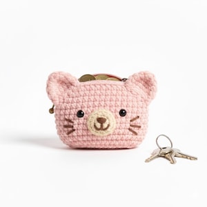 May include: A pink crocheted coin purse shaped like a cat's face, with black eyes, a tan nose, and whiskers. The purse is filled with coins and has a zipper closure. A set of keys rests beside the purse.