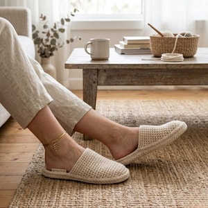 Peut inclure: Des chaussons beiges en tissu tissé avec un bout arrondi. Les chaussons sont portés sur un tapis en jute. Une chevillère dorée en forme de feuille est visible. Le texte "ON YOU HAIR" est imprimé sur la semelle.
