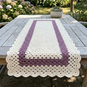 Peut inclure: Chemin de table rectangulaire en crochet crème et violet. Le centre est crème, bordé de violet et orné d'une bordure festonnée. Posé sur une table en bois, il est mis en valeur dans un jardin.