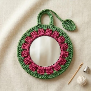 May include: A round, hand-crocheted mirror with a green border and a pink floral design around the mirror. The mirror has a green handle and a leaf-shaped accent. A needle, thimble, and ball of yarn are next to the mirror.