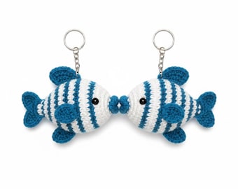 May include: Two crocheted fish keyrings, each with a white body and blue stripes, fins, and tail. The fish are kissing, with their lips touching. Each keyring has a silver ring attached to the top.
