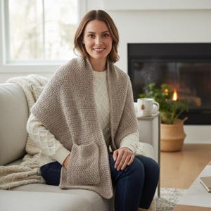 May include: A beige crocheted shawl with a pocket, worn over a white sweater and blue jeans. The shawl is draped over the shoulders and has a pocket on the front. The woman is sitting on a gray couch.