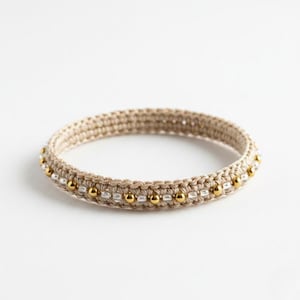 Puede incluir: Una pulsera de ganchillo beige con cuentas doradas y plateadas. La pulsera es circular y parece hecha a mano. Las cuentas están espaciadas uniformemente alrededor de la pulsera, añadiendo un toque de elegancia.