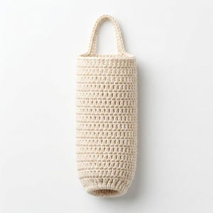 May include: A cream-colored, crocheted bag with a handle. The bag is cylindrical with a rounded bottom and a looped handle at the top. The crochet stitches are visible, creating a textured surface. The bag is designed to hold items.
