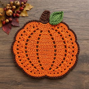 May include: A pumpkin-shaped crochet doily featuring a bright orange center with an openwork lattice pattern, a dark brown stem, and a single green leaf. The piece is edged with a dark brown border, creating a distinct pumpkin silhouette.